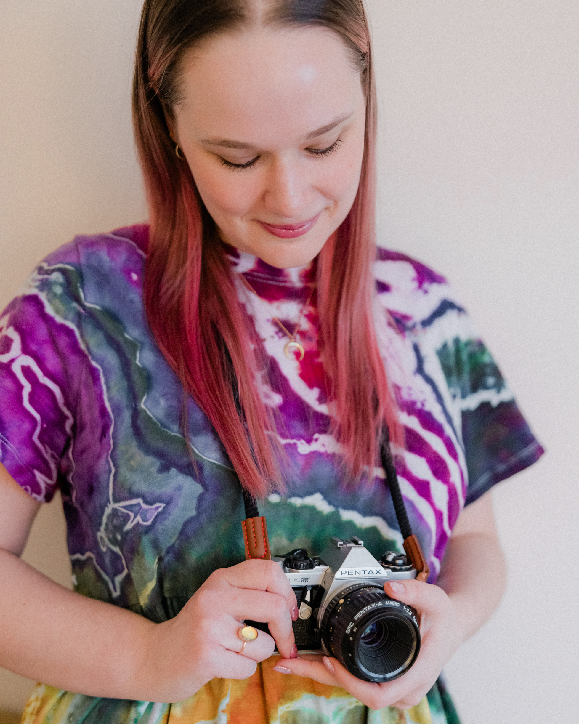 Amber, wearing a tie-dye dress, looking down and smiling at the vintage camera she's holding against a plain background