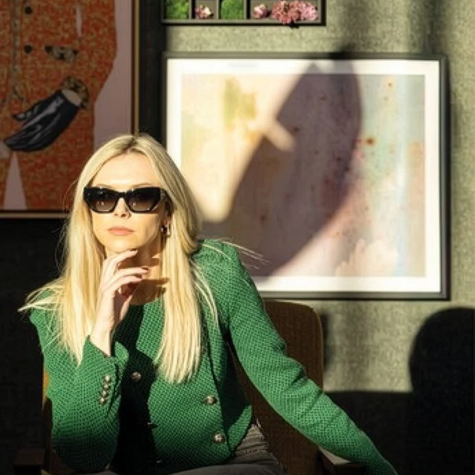 A blonde woman wearing black sunglasses and a textured green jacket sits in a chair in a sunlit room with a framed art print by Amber Lauder behind her.