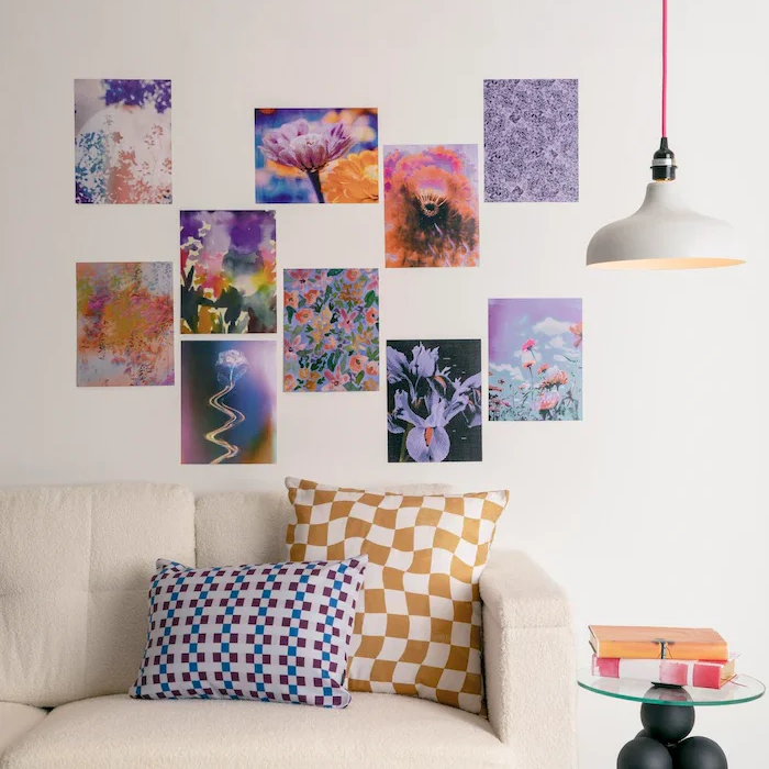 A styled living room scene showing a wall arrangement of multiple colorful abstract and floral art prints, including Amber Lauder's artwork, above a cream sofa with patterned pillows, plus a hanging pendant lamp and side table.