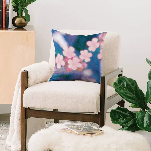 A square throw pillow with a pink floral pattern on a blue and purple background, titled Delicate by Amber Lauder, styled on a cream-colored armchair in a cozy room setting.