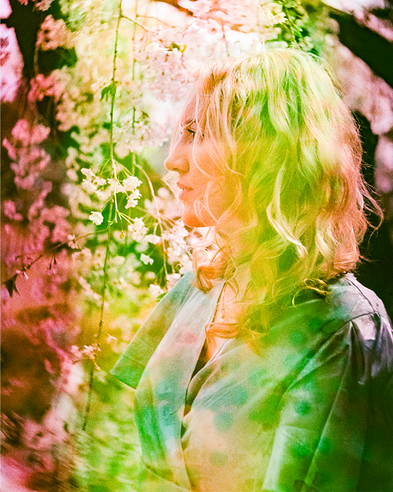 Hannah Telle in profile surrounded by soft-focus pink blossoms and overlaid with a vibrant green, pink and yellow light leak effect.